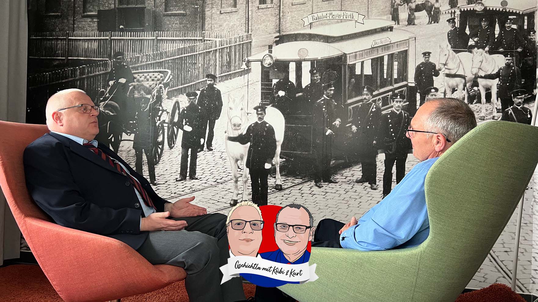 Gschichtla mit Kübi & Kurt: Beide sitzen vor einem historischen Foto im Betriebshof Heinrich-Alfes-Straße